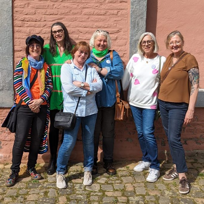 Sechs Frauen stehen vor einer mauerartigen Oberfläche, lächeln und posieren.