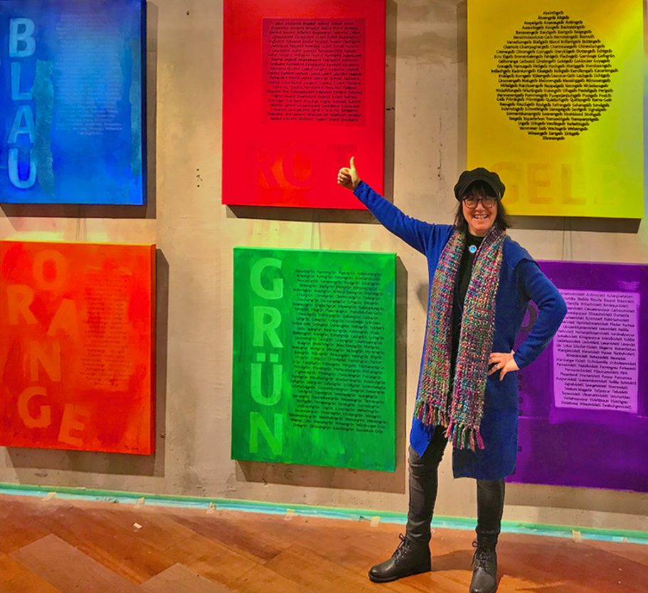 Person in blauer Jacke zeigt auf farbige Kunstwerke an der Wand, die Farben übereinander anordnen.