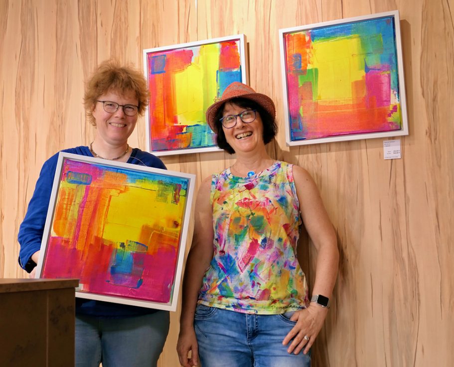 Zwei Frauen stehen neben bunten, abstrakten Bildern an einer Holzwand.