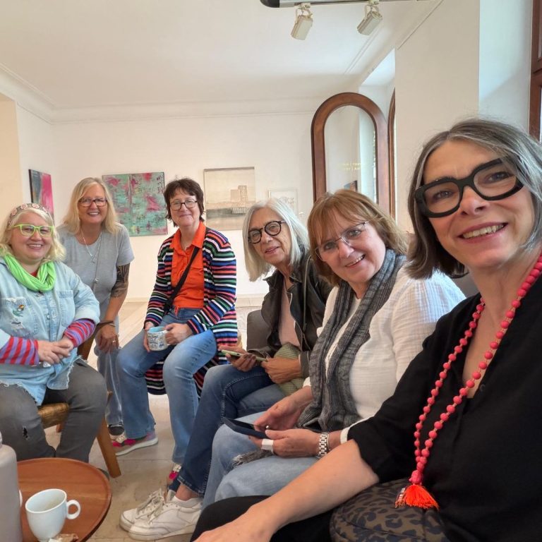 Sechs Frauen lächeln in einem hellen Raum, sitzen zusammen und unterhalten sich.