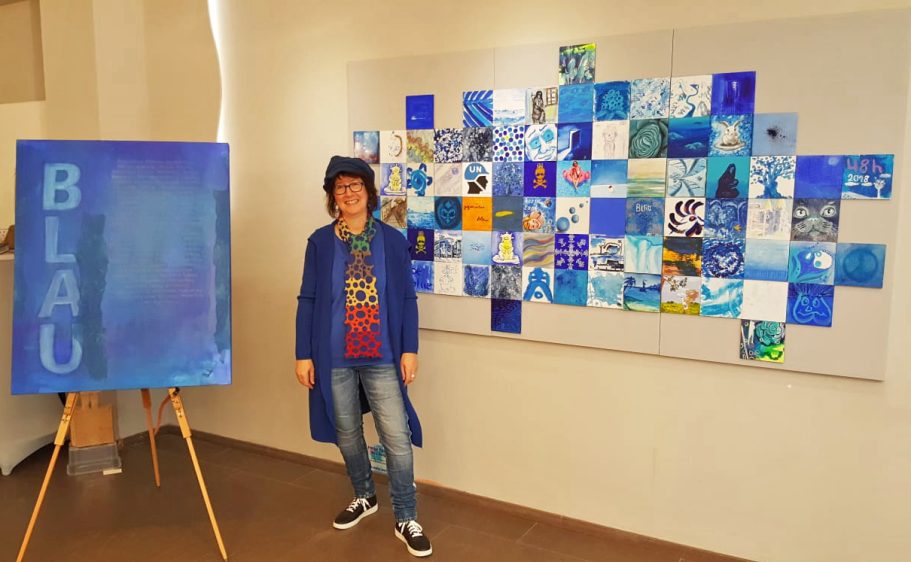 Künstler in blauer Kleidung neben einer Wand mit blauen Kunstwerken und einem Gemälde.