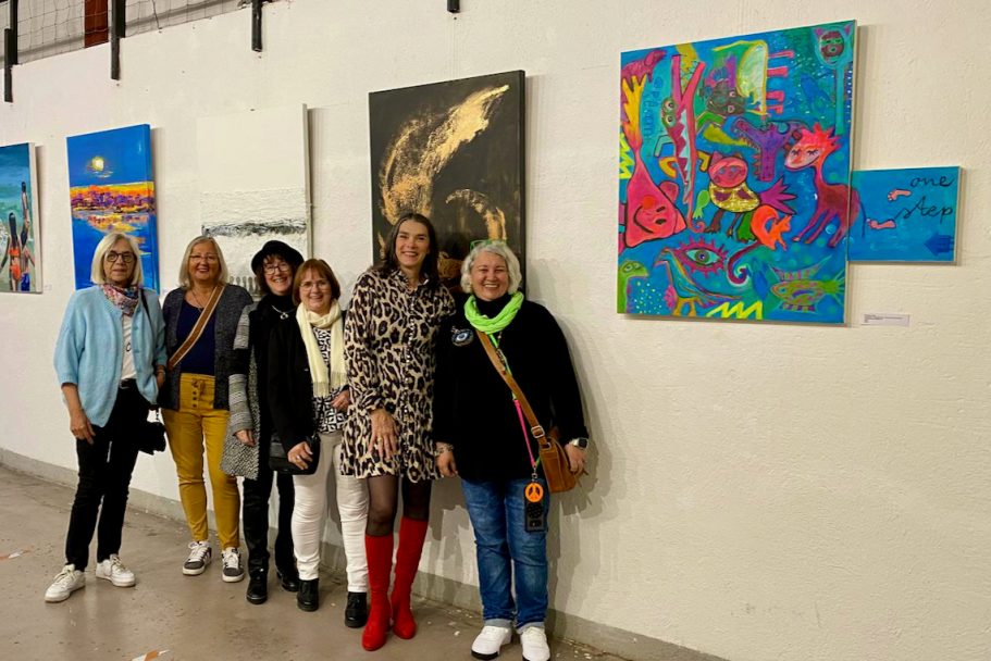 Sechs Frauen stehen vor bunter Kunst an einer Wand in einer Galerie.