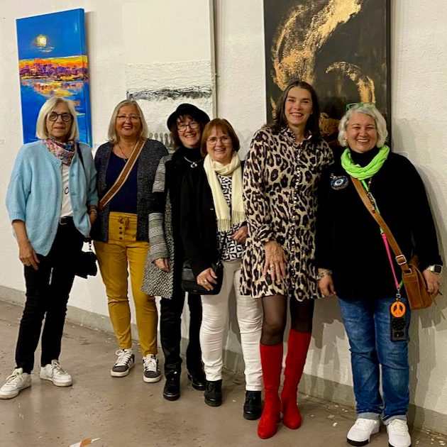 Sechs Frauen stehen lächelnd vor einer Kunstausstellung, einige tragen farbenfrohe Outfits.