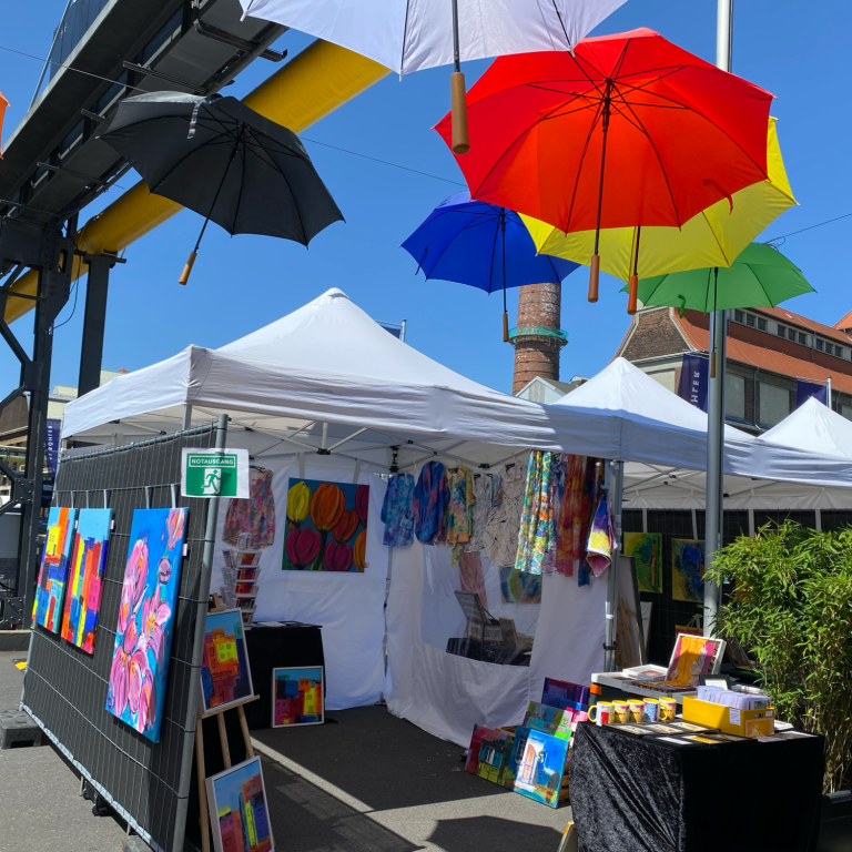 Bunte Regenschirme hängen über einem Marktstand mit Kunstwerken und Farben.