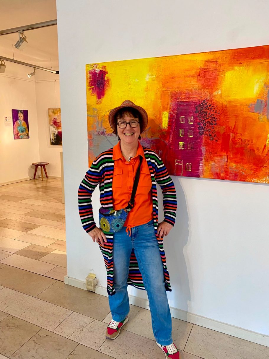 Frau in gestreiftem Mantel und orangefarbenem Shirt steht vor farbenfrohem Kunstwerk.