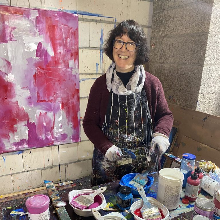 Frau mit Brille und Schürze lächelt vor einer Farbpalette und Malutensilien.