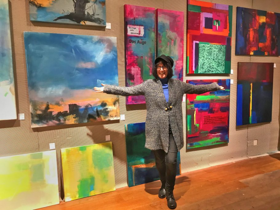 Frau in grauem Mantel steht vor bunten, abstrakten Gemälden in einer Galerie.