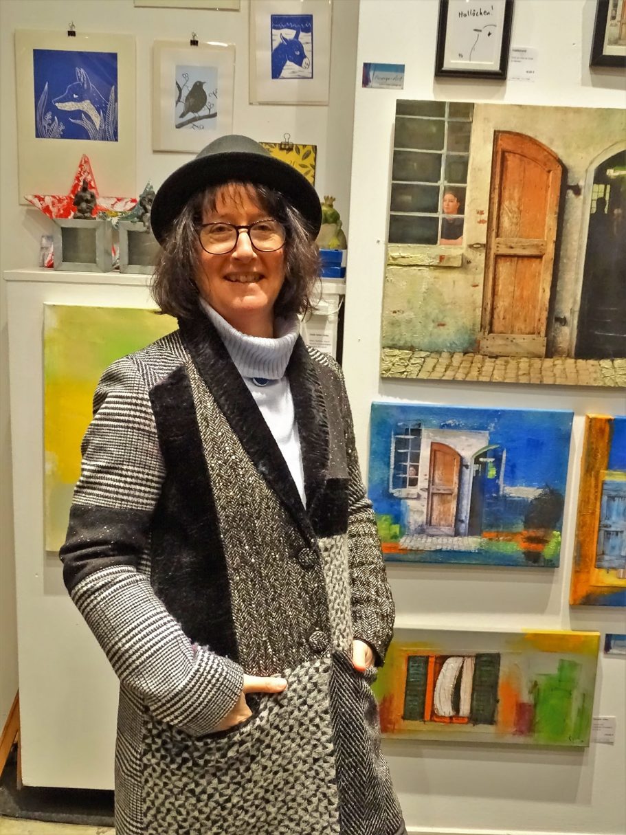 Frau in Hut und grauem Mantel steht vor einer Galerie mit bunten Kunstwerken.