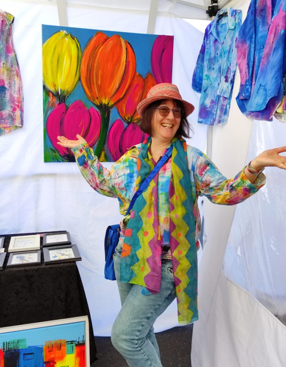 Frau mit buntem Outfit und Hut posiert vor einer Kunstinstallation mit Blumenmotiven.