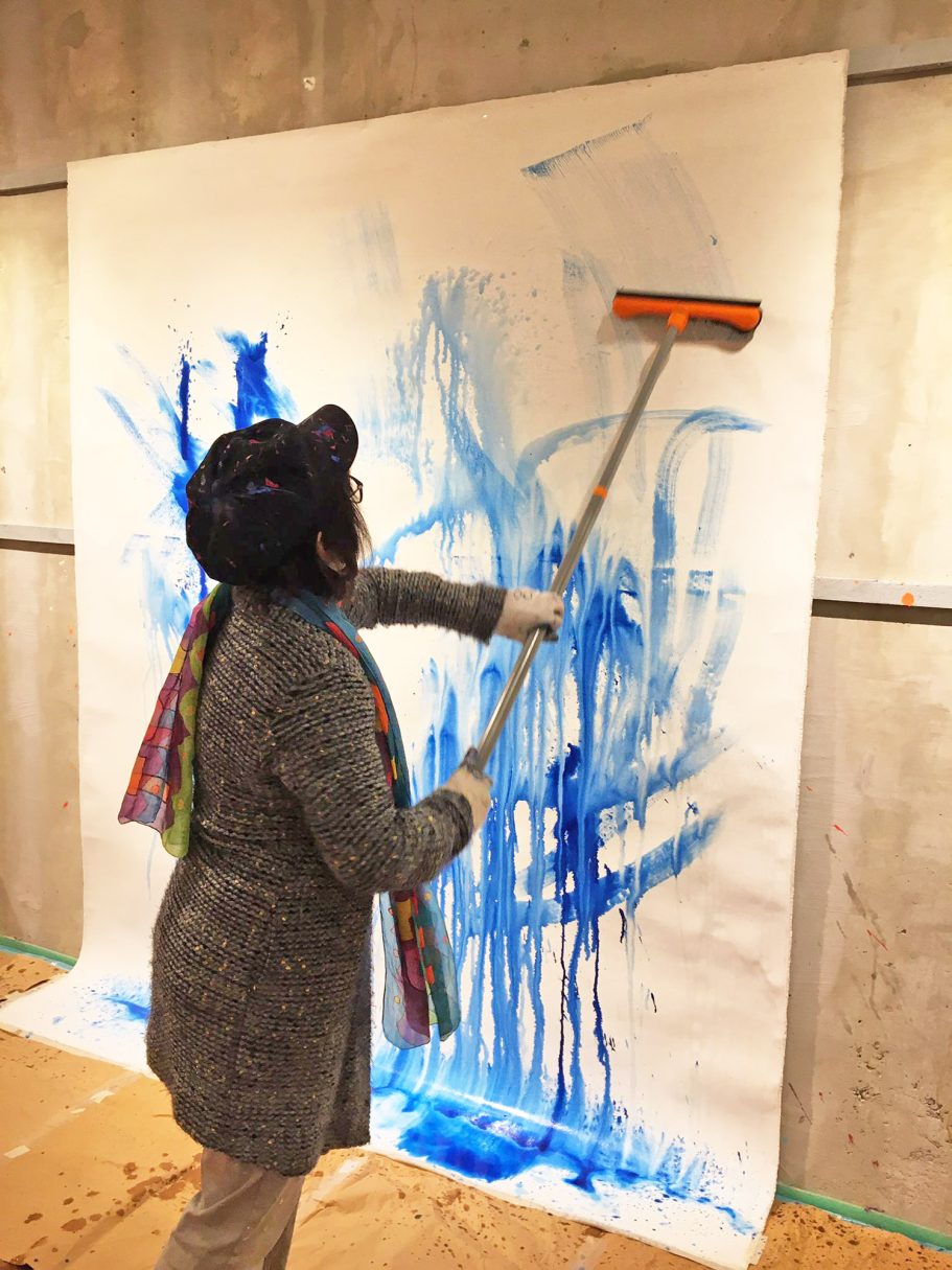 Eine Person streicht mit einer Walze blaue Farbe auf eine große Leinwand.