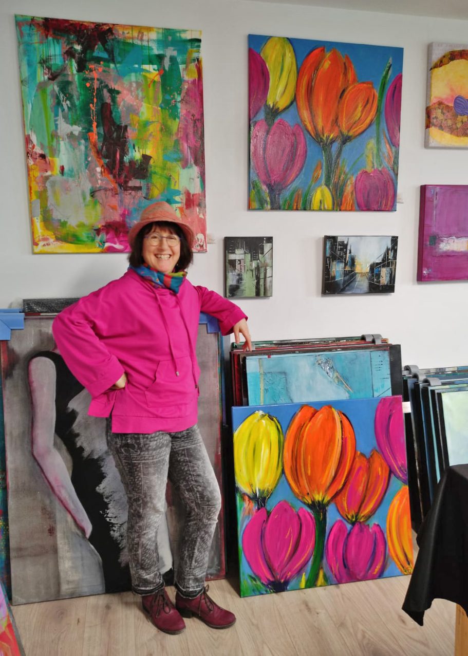 Frau in pinker Jacke steht neben bunten Blumenbildern in einem Atelier.