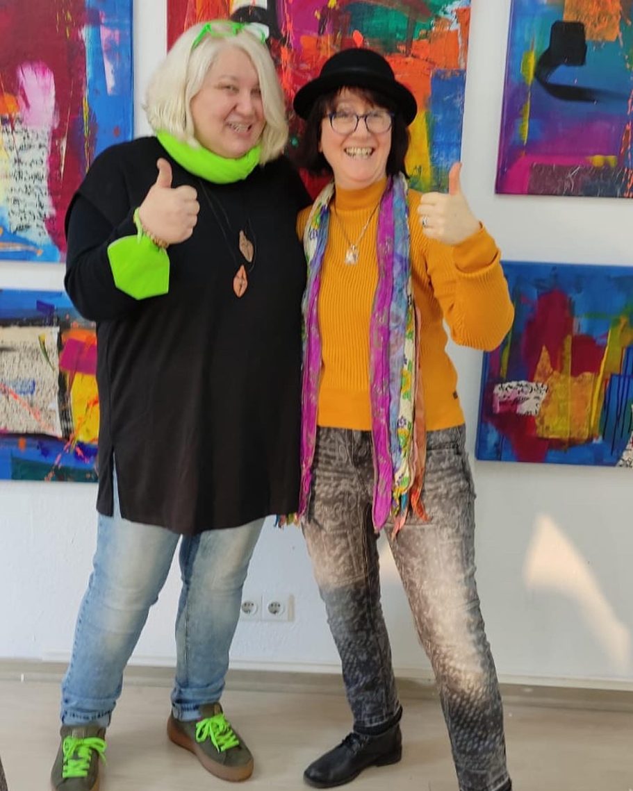 Zwei Personen mit Daumen hoch, vor bunten abstrakten Gemälden.