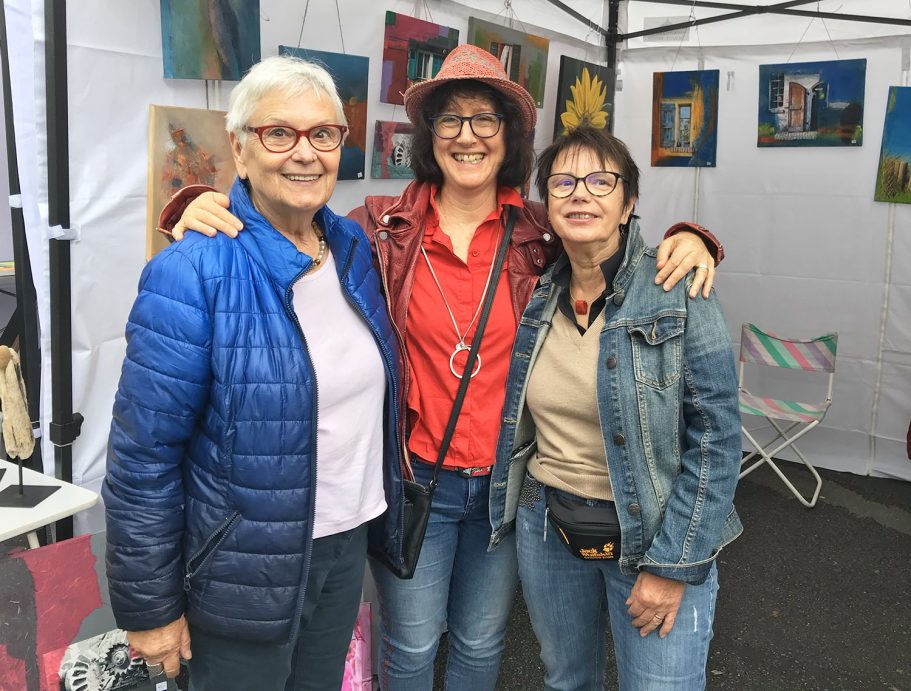 Drei Frauen lächeln fröhlich in einem Kunstzelt, umgeben von bunten Bildern.